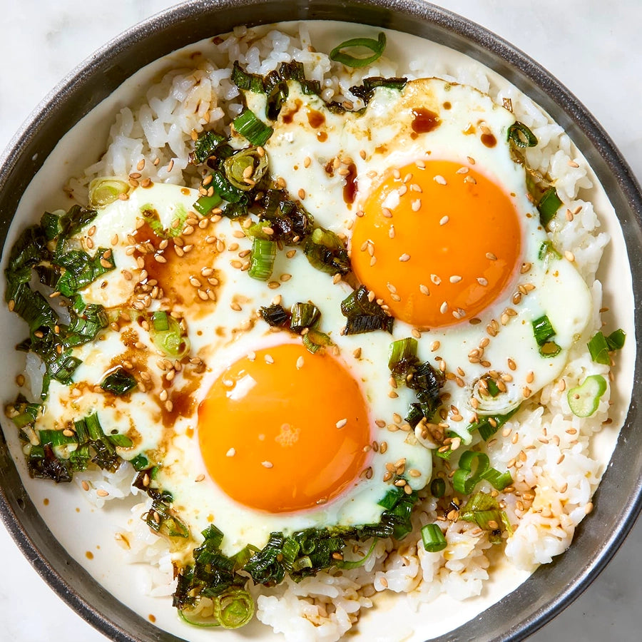 Bowl of rice with two fried eggs, green onions, and sesame seeds on a white background