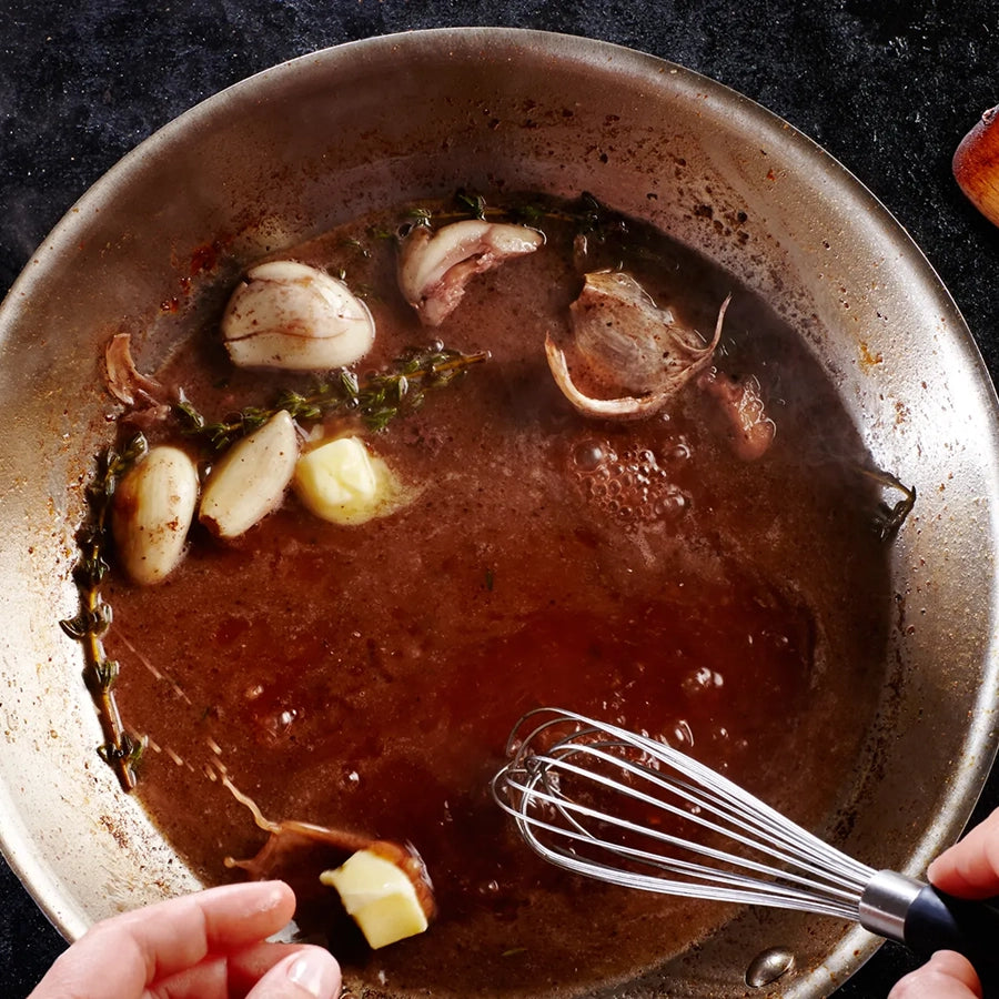 Brown sauce with garlic cloves and herbs in a pan with a whisk