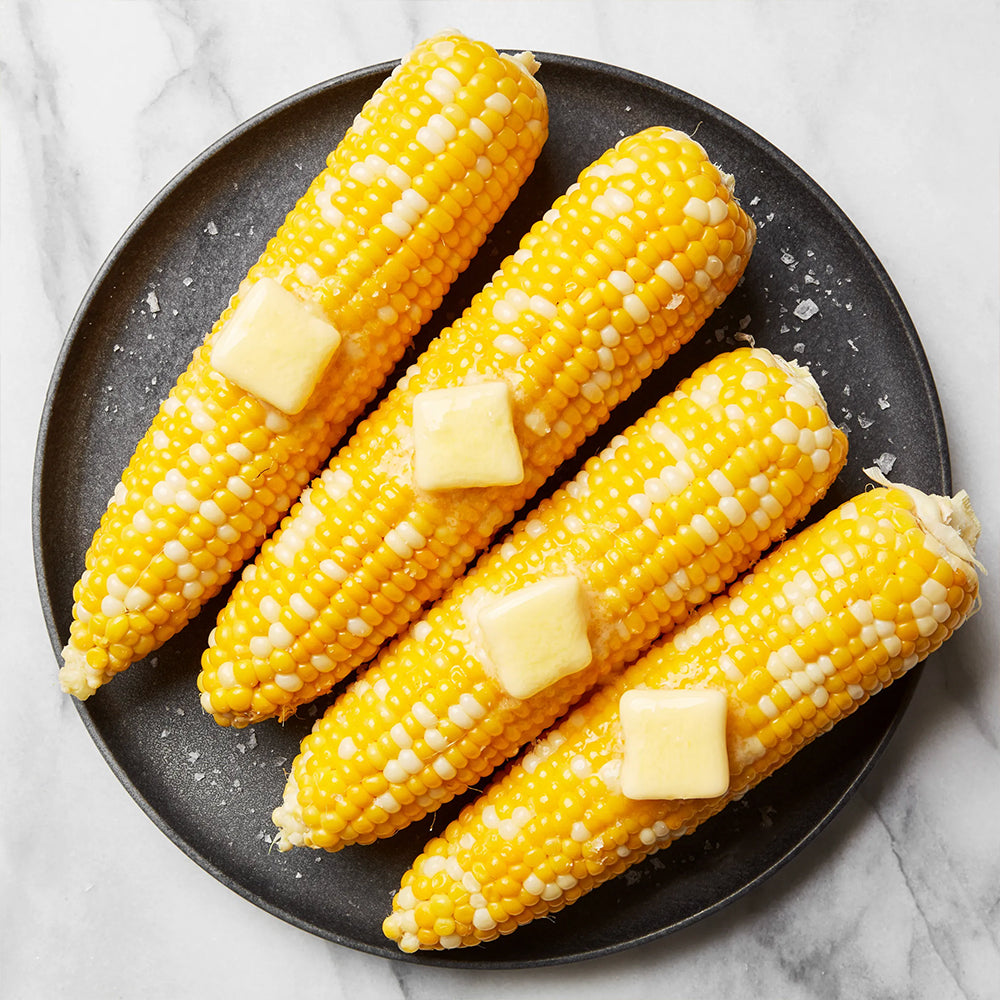 Four ears of corn on a black plate with butter on top, on a marble surface.