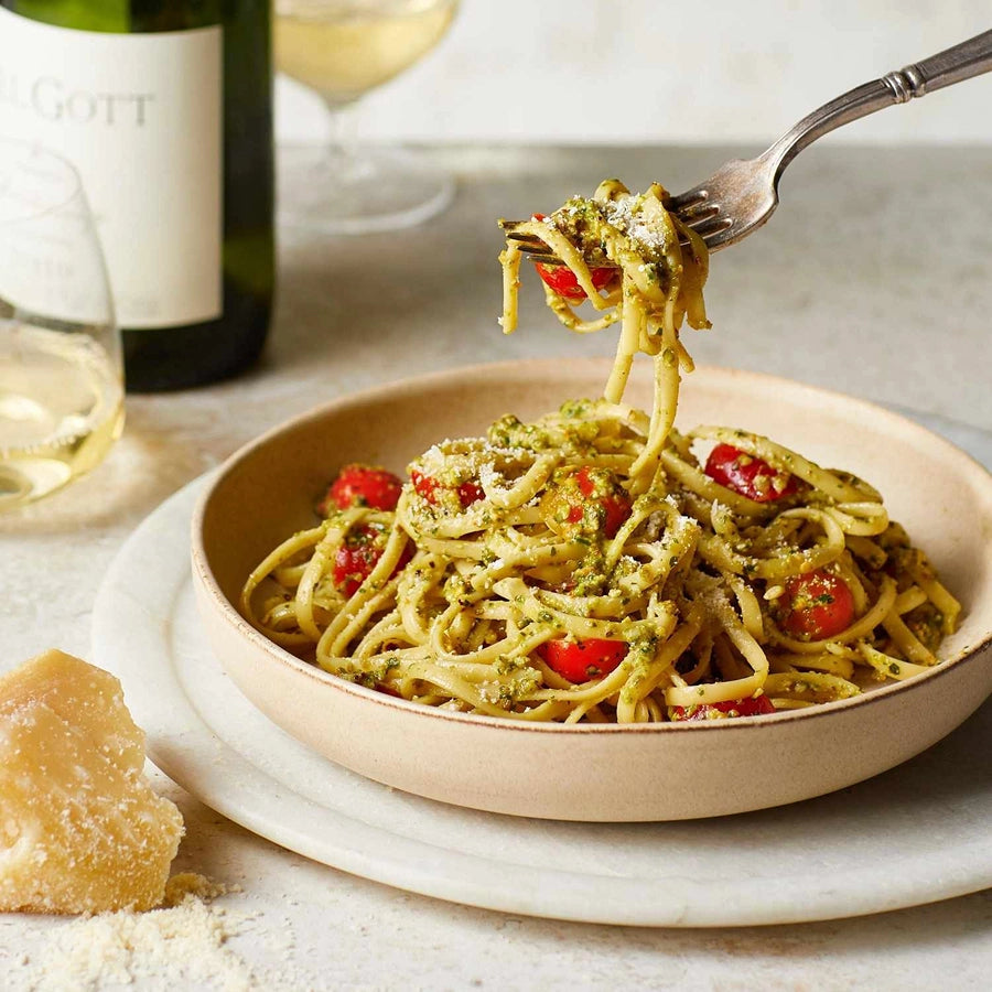 Pasta dish with pesto and tomatoes on a plate, served with a bottle of wine and glasses in the background.